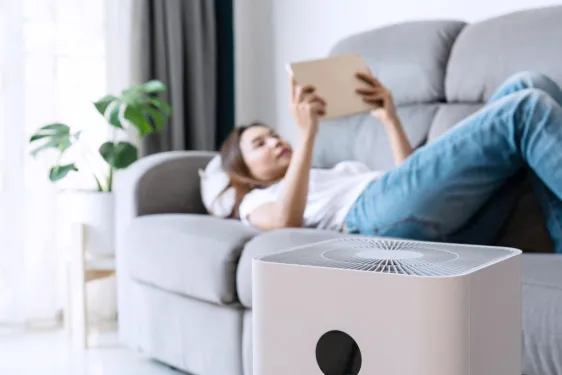 Picture of woman laying on couch and reading with an air purifier in the foreground.